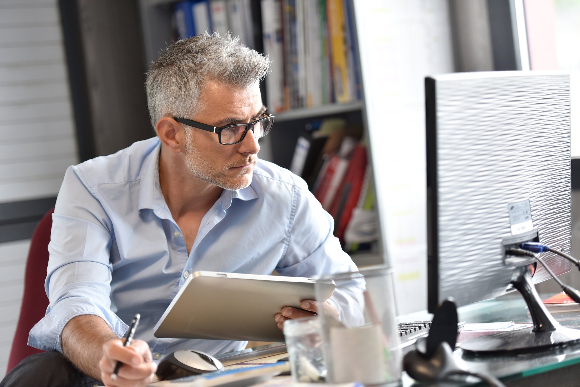 Middlle age business man with glasses and grey hair, holding a tablet and looking at laptop. 
